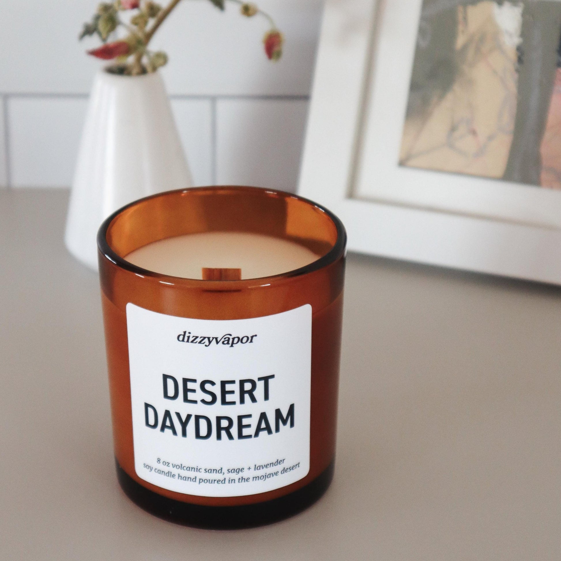 A brown amber glass jar candle with a white label that reads 'desert daydream'. It is placed on a flat surface with a white object in the background.